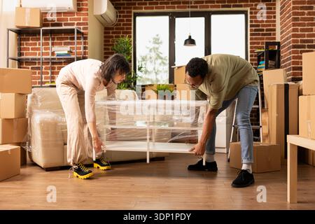 Marito afroamericano e moglie caucasica che trasportano un tavolino da caffè avvolto all'interno di una nuova parete di mattoni. Scrivania multietnica per coppie sposate e arredamento accogliente il giorno del trasloco. Foto Stock