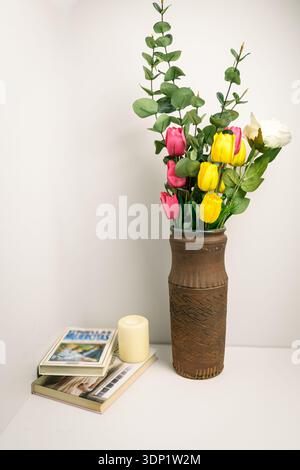 Tulipani colorati ed eucalipto in un vaso in terracotta rustico con libri e candele su uno scaffale bianco, natura morta minimalista con spazio per le copie. Foto Stock