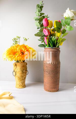 Interni minimali con due vasi: Alto vaso in terracotta con tulipani, rosa ed eucalipto e vaso rustico con margherite gerbera arancioni su tabl bianco Foto Stock