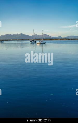 Le barche a vela galleggiano pacificamente su acque calme, riflettendo il cielo, con montagne in lontananza, l'Irlanda. Foto Stock