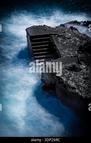 Foto a lunga esposizione per catturare una struttura costiera in cemento scuro. L'uso di una velocità dell'otturatore ridotta crea l'effetto setoso dell'acqua, contrastando il wi Foto Stock