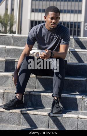 Uomo afroamericano seduto su passi concreti in abbigliamento sportivo con cuffie e orologio da controllo Foto Stock