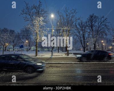 Le auto sfocate attraversano forti nevicate su un viale bagnato a Belgrado, Serbia, di notte, illuminato da semafori e cavi del tram. Guida invernale, slus Foto Stock