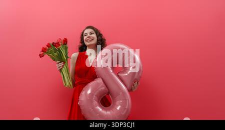 Giovane donna felice in un vivace abito rosso racchiude un bouquet di tulipani rossi e un palloncino rosa brillante numero 8, che irradia positività e potere contro Foto Stock