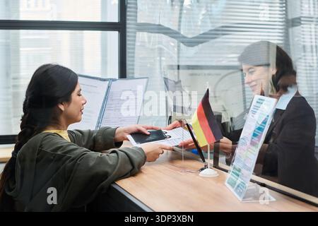 Giovane donna latina adulta che presenta passaporto e documenti a una donna caucasica di mezza età dietro un bicchiere al banco del centro visti, entrambi interagiscono con i documenti cartacei e con la bandiera tedesca visibile sulla scrivania Foto Stock