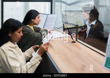 Due giovani donne ispaniche adulte che presentano documenti al banco del centro visti, interagiscono con una donna caucasica di mezza età dietro un divisorio di vetro, tenendo in mano documenti di identificazione Foto Stock