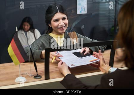 Giovane donna ispanica sorridente mentre consegna il passaporto e il modulo di richiesta del visto a una donna caucasica di mezza età dietro il bancone del centro visti, bandiera tedesca alla reception Foto Stock