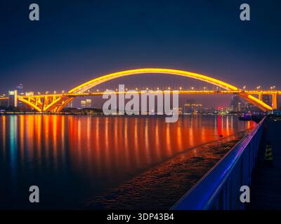 Ponte Lupu illuminato che attraversa il fiume Huangpu di notte, con sentieri luminosi e riflessi sullo skyline della città. Foto Stock