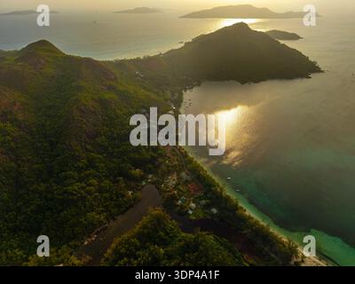 La vista aerea della luce del sole dorata bacia le acque turchesi che circondano lussureggianti colline verdi e il villaggio costiero di Baie Ste Anne, Praslin, Seychel Foto Stock