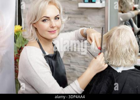 Donna parrucchiere sorridente seduta e tagliata i capelli di una donna bionda. Foto Stock