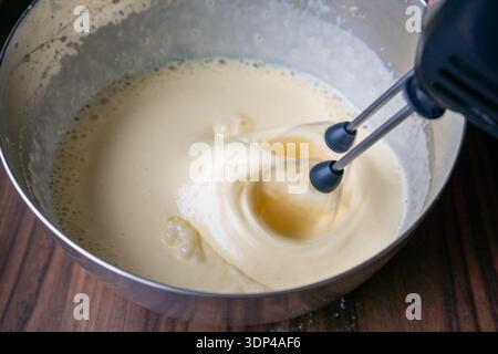 Vista ravvicinata dell'impasto liquido per torte miscelato con un mixer manuale elettrico all'interno di un recipiente di metallo. Foto Stock