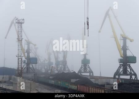 Porto industriale con gru da carico di grandi dimensioni che operano in condizioni di forte nebbia. Sulla piattaforma di carico sono visibili pile di treni per carbone e merci, creando un dramma Foto Stock