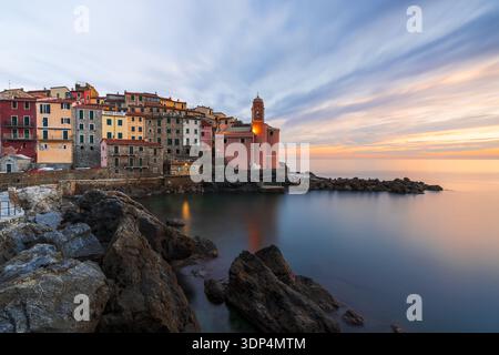 Vista degli edifici color pastello aggrappati alla costa rocciosa, una chiesa immersa nel soffice bagliore del tramonto che si affaccia sul tranquillo mare di Tellaro, li Foto Stock