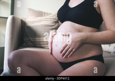 Donna incinta che abbraccia la pancia in un ambiente confortevole Foto Stock