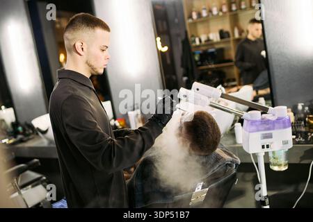 Giovane uomo caucasico adulto che indossa guanti che esegue un trattamento al vapore facciale su giovane adulto uomo caucasico reclinato su una sedia da barbiere con attrezzature professionali per la cura della pelle visibili Foto Stock