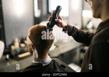 Giovane uomo adulto caucasico seduto su una sedia da barbiere che riceve un taglio di capelli dal giovane barbiere adulto caucasico che utilizza un regolacapelli elettrico, entrambi focalizzati sul processo di cura del corpo Foto Stock
