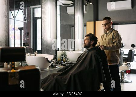 Uomo di mezza età caucasico seduto sulla sedia del barbiere, che viene tagliato di capelli da un giovane uomo caucasico adulto in piedi dietro di lui usando mantello da barbiere e strumenti professionali vicino a specchi e lavandini Foto Stock