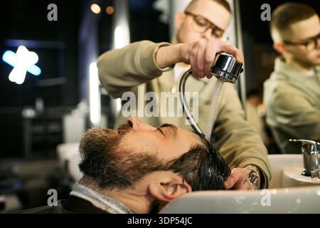 Giovane uomo adulto caucasico che lava i capelli di un uomo caucasico di mezza età al lavandino del barbiere, concentrandosi sul processo di lavaggio professionale dei capelli con acqua che scorre sopra la testa dei clienti Foto Stock