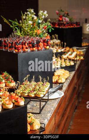 Elegante tavolo da pranzo con vibranti bicchieri da shot pieni di bacca, mini crostate assortite e tartine gourmet su riser neri Foto Stock