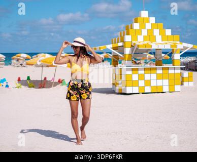 Goditi una giornata pittoresca a Miami Beach, una donna in abbigliamento da spiaggia alla moda passeggia lungo la spiaggia di sabbia bianca. Ombrelloni colorati offrono ombra, esaltando l'atmosfera vibrante di questa vacanza al mare. Foto Stock