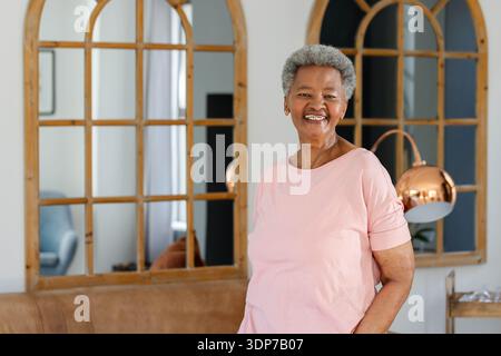 Donna afroamericana anziana in piedi e sorridente a casa, vicino a specchi di legno ad arco, spazio fotocopie Foto Stock