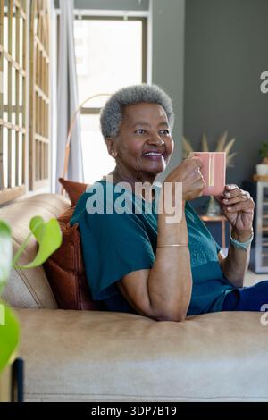 Donna afroamericana anziana che indossa la parte superiore color verde acqua seduto sul divano di casa tenendo in mano una tazza rosa Foto Stock