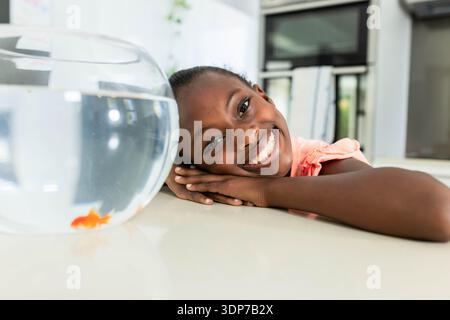 Bambina afroamericana che riposa la testa su un piano lucido in cucina guardando la ciotola dei pesci rossi Foto Stock