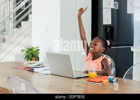 Giovane donna afroamericana che indossa la parte superiore rosa seduta a un tavolo di casa usando un computer portatile, alzando la mano Foto Stock