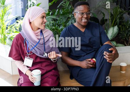 Diversi colleghi in scrub seduti sulla panchina in sala colazione con stetoscopio che tiene caffè e mela Foto Stock