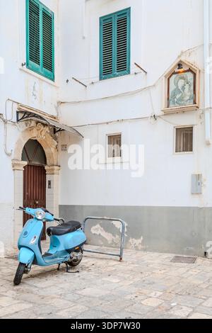 Uno scooter Vespa azzurro è parcheggiato fuori da un edificio imbiancato a calce con persiane verdi in un vicolo italiano. a Monopoli Foto Stock