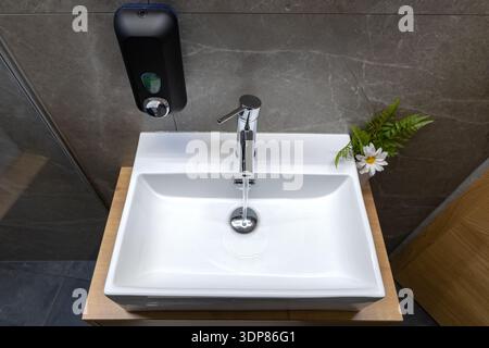 Un moderno lavandino in ceramica bianca con rubinetto cromato lucido è installato in un bagno moderno con pareti grigie, un dispenser di sapone e una piccola pianta decorativa Foto Stock