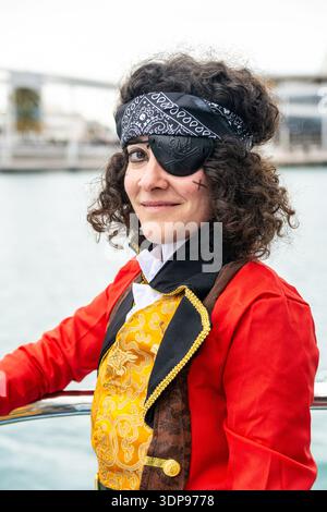 Donna sorridente in un vivace costume da pirata con patch per gli occhi, bandana e giacca rossa in un vivace carnevale sul lungomare, capelli ricci e trucco natalizio Foto Stock