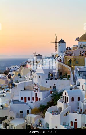 I turisti si riuniscono a Oia, Santorini, in Grecia, per assistere all'iconico tramonto sul Mar Egeo, circondato da tradizionali edifici bianchi e dal vento Foto Stock