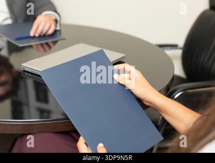 Due persone siedono ad un tavolo rotondo in un ufficio. Una persona tiene una cartella mentre l'altra è seduta con un notebook. Probabilmente stanno discutendo di un importante busin Foto Stock