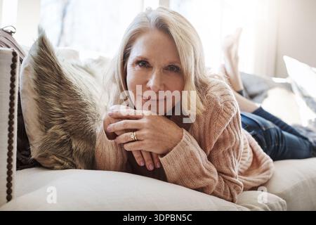 Accogliente, ritratto e sorriso di una donna sul divano nel soggiorno di casa per rilassarmi o fare una pausa. Pausa, felice e rilassati con una persona matura sdraiata Foto Stock