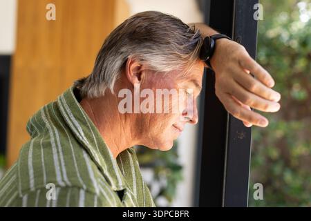 Uomo maturo che si appoggia sulla cornice della finestra scura a casa, che guarda verso il basso, indossa una camicia verde, guarda Foto Stock