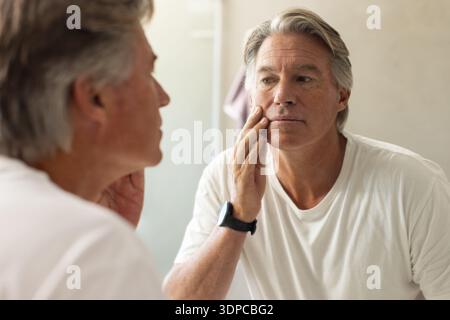 Uomo anziano in piedi allo specchio del bagno che tocca la guancia indossando T-shirt e orologio da polso bianchi Foto Stock