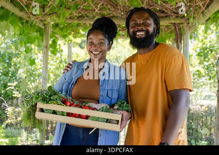 Coppia che porta una cassa di pomodori e verdure sotto la pergola della vite in giardino in denim e camicie arancioni Foto Stock