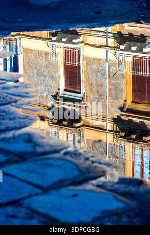 Riflesso di edifici residenziali con balconi e persiane in una pozza d'acqua su pavimentazione in pietra nel centro storico di Catania, Italia. Foto Stock