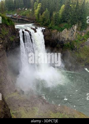 Maestosa cascata che scende giù dalle scogliere rocciose circondate da lussureggiante vegetazione e nebbia, con un ponte in legno e edifici visibili sullo sfondo Foto Stock