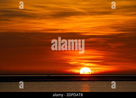 Tramonto vibrante su un paesaggio costiero dove un lontano parco eolico si staglia contro un cielo brillante e ardente Foto Stock