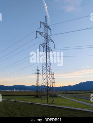 Linee elettriche e torri di trasmissione si ergono alte in un'area rurale con montagne sullo sfondo, mostrando infrastrutture elettriche al crepuscolo. Foto Stock