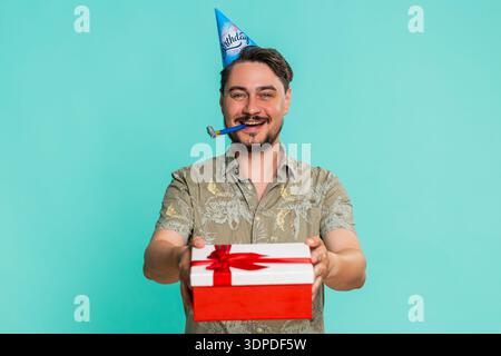 Un uomo di mezza età che indossa un cappello festoso sorride calorosamente, fa felicemente un regalo di compleanno, celebrando chiaramente la generosità e la gentilezza. Ragazzo adulto isolato su sfondo blu che si gode una festa gioiosa Foto Stock