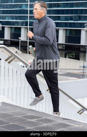 Uomo di mezza età che sale le scale urbane in un abbigliamento sportivo che tiene in mano una bottiglia testurizzata, indossa auricolari, guarda Foto Stock