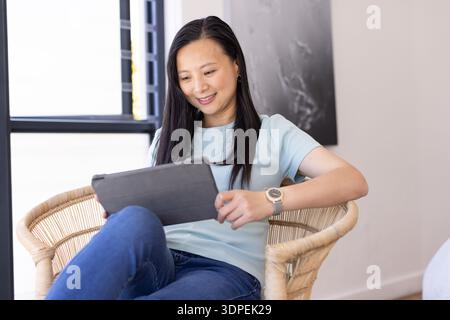 Donna asiatica seduta in una poltrona di rattan intrecciata vicino alla finestra che tiene un tablet, indossando un orologio da polso Foto Stock