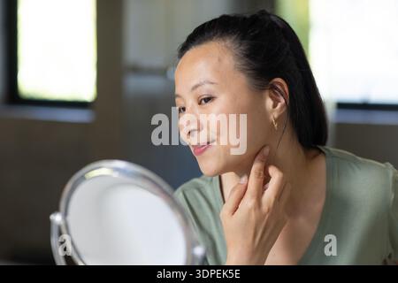 Donna asiatica alla vanità che controlla la pelle nello specchio sotto la luce della finestra, indossando cerchi d'oro con scollo a V verde Foto Stock