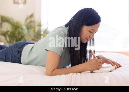 Donna asiatica sdraiata sullo stomaco scrivendo in un taccuino con matita sul letto che indossa la parte superiore verde chiaro Foto Stock