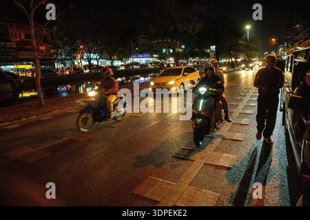 Una dinamica scena notturna in una strada trafficata a Chiang mai, Thailandia. Motociclette, automobili e un pedone si spostano lungo una strada accanto a un canale, illuminata da Foto Stock