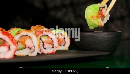 I panini di sushi sono posizionati su un piatto nero mentre un pezzo viene raccolto con le bacchette e immerso nella salsa di soia Foto Stock