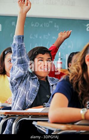 Un ragazzo latino si siede a una scrivania in una classe con la mano alzata, desideroso di partecipare alla lezione mentre i compagni di classe prestano attenzione. L'impostazione è foc Foto Stock